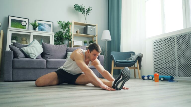 Man stretching on living room floor doing a home workout with dumbbells and protein shaker nearby